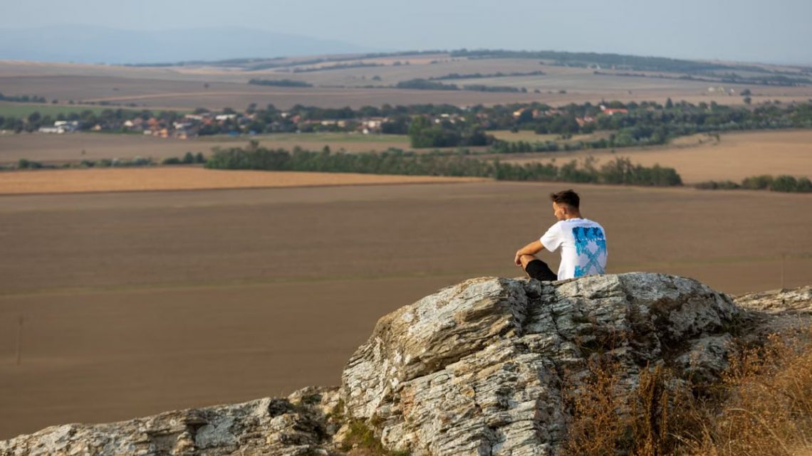 A man sits on a rock overlooking the vast countryside in a serene outdoor setting.